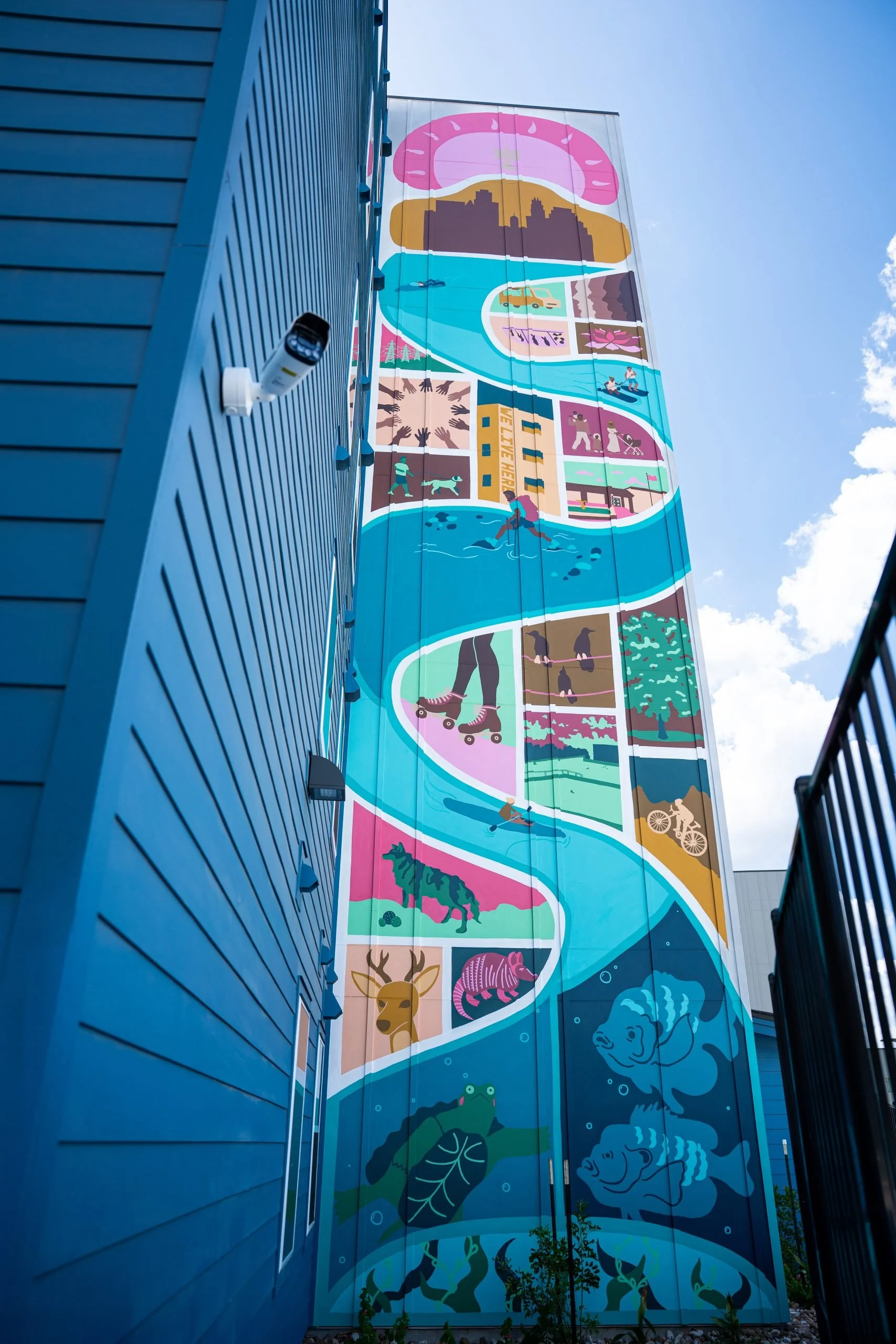 Colorful mural on a tall building featuring various animals, outdoor activities, and scenic landscapes, with a blue sky in the background.