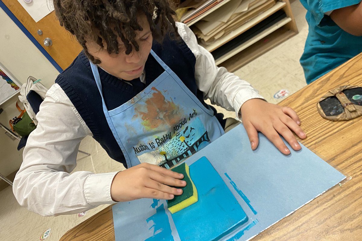 A young boy is sponging a piece of blue artwork on paper in a classroom.