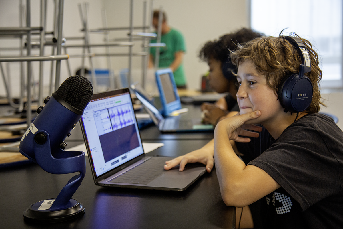 A young boy wearing headphones sitting at a table with a laptop and microphone, recording or editing audio, in a classroom or studio with other students working in the background.