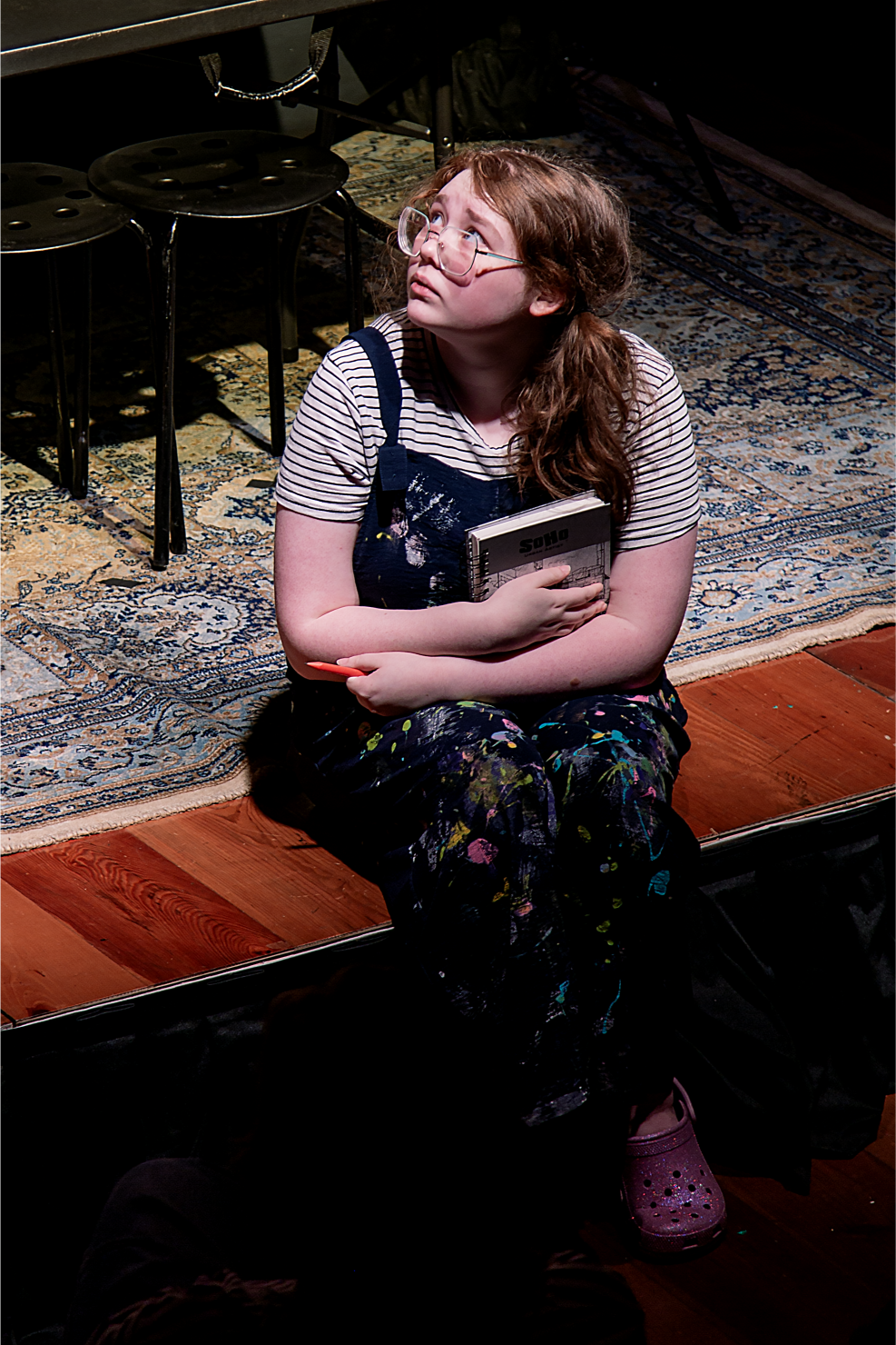 A girl with glasses, red hair, and wearing a striped shirt and paint-splattered overalls sitting on the stage floor, holding a notebook and looking upward with a worried or thoughtful expression.