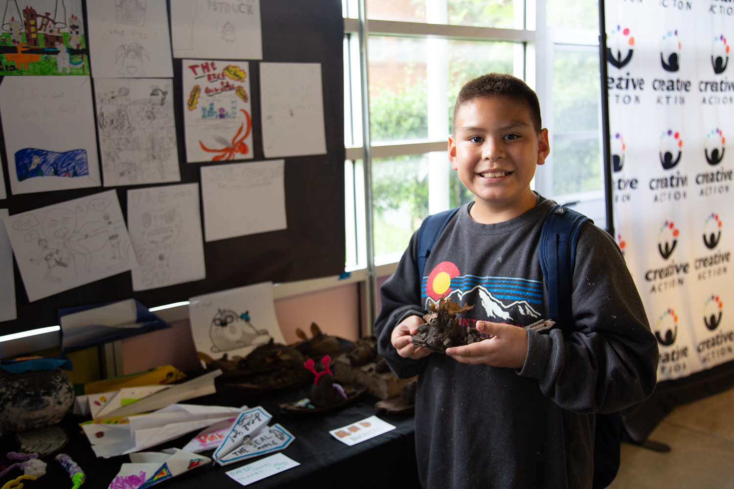 A young boy smiling at the camera while holding a piece of clay artwork. Behind him are art projects and drawings on display, with a window to the outside showing trees. The classroom setting is lively with colorful artwork.