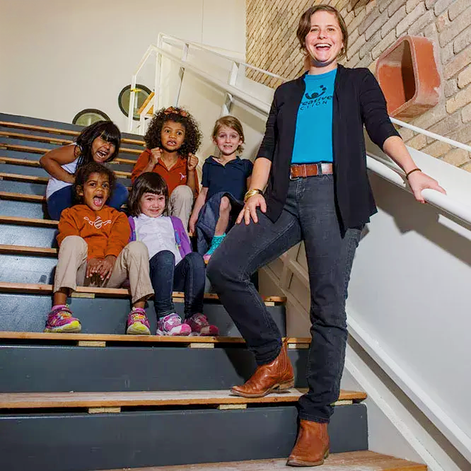 Founder Karen LaShelle and five children sitting on stairs, posing for a photo in an indoor setting with a brick wall and a calendar or notice board in the background.