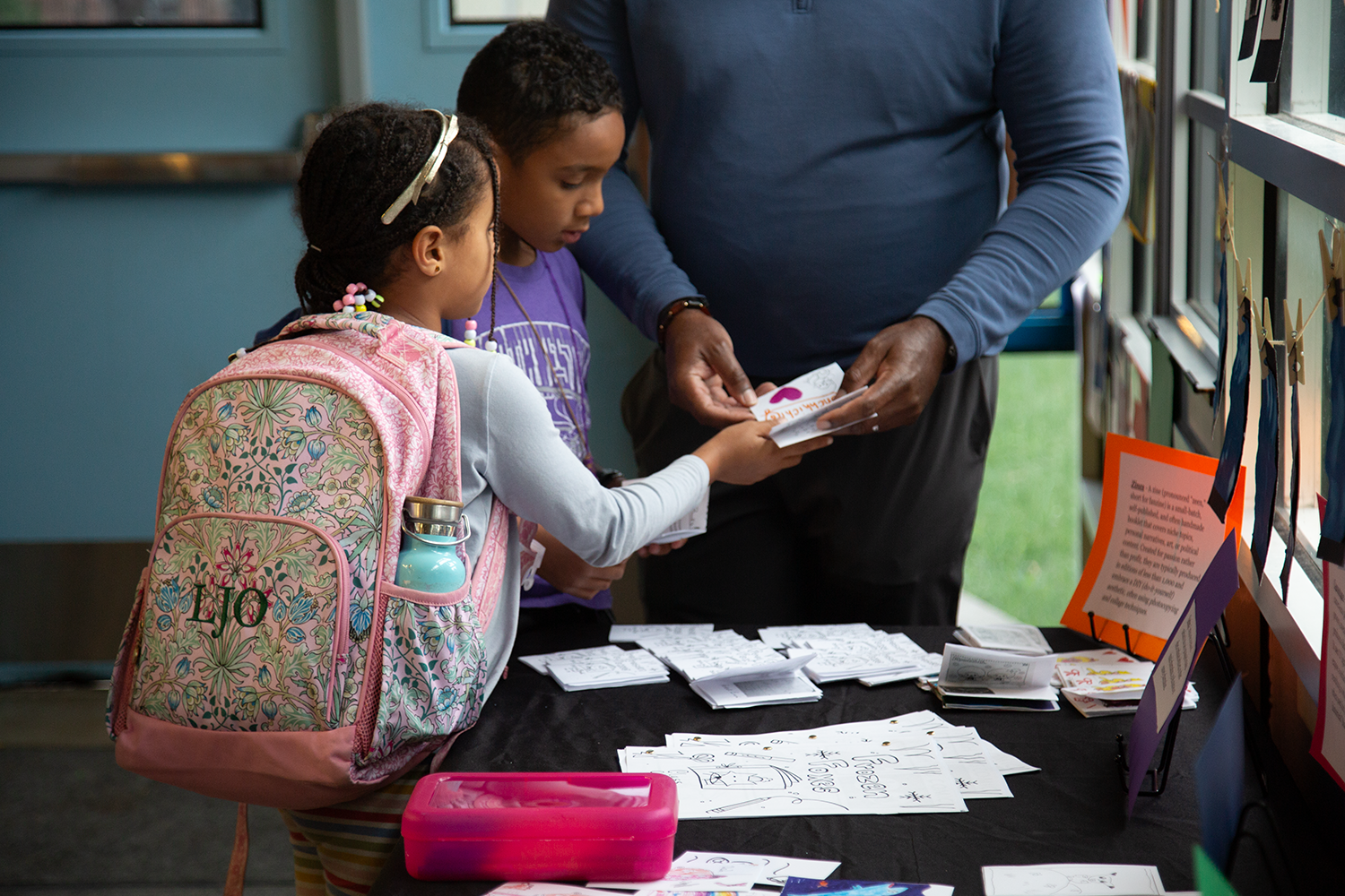A man and two young girls are at a table, looking at projects during an after school showcase.