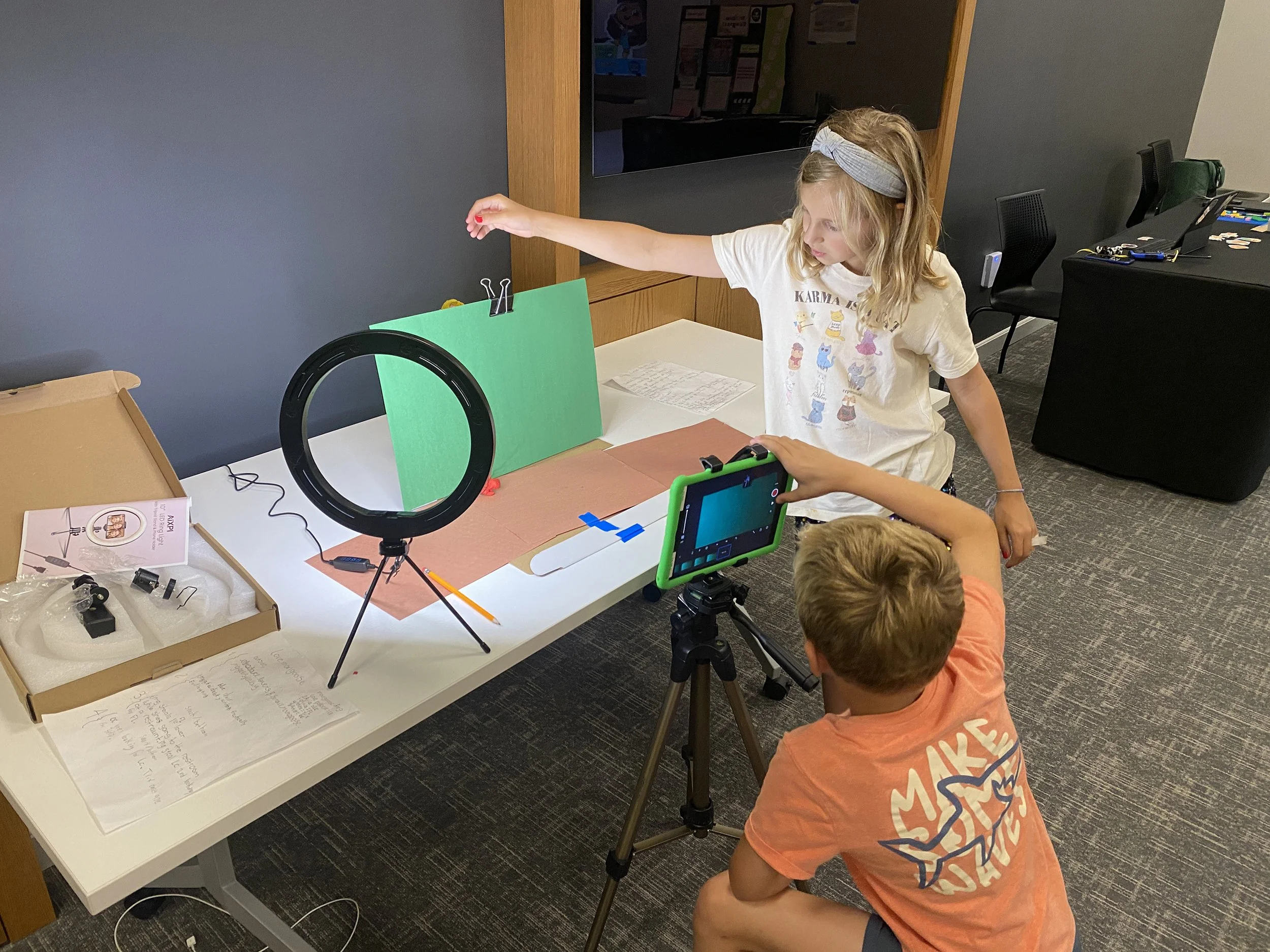 A young girl with blonde hair tied up with a gray headband is reaching her arm across a table in a classroom or activity room. She is wearing a white T-shirt with colorful illustrations. The table has various objects, including a green circular track, papers, a tablet, and a ring light. Several children are partially visible, and there's a black desk with items on it in the background.