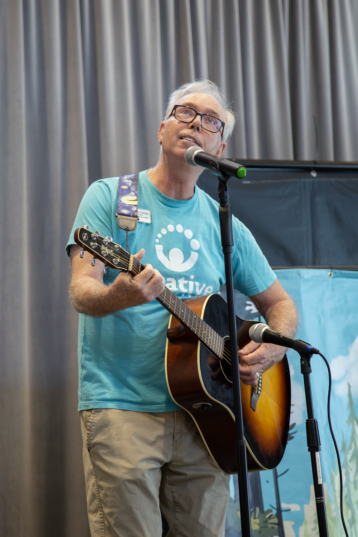 An older man with gray hair, glasses, and a blue T-shirt playing an acoustic guitar and singing into a microphone on a stage with a curtain backdrop.