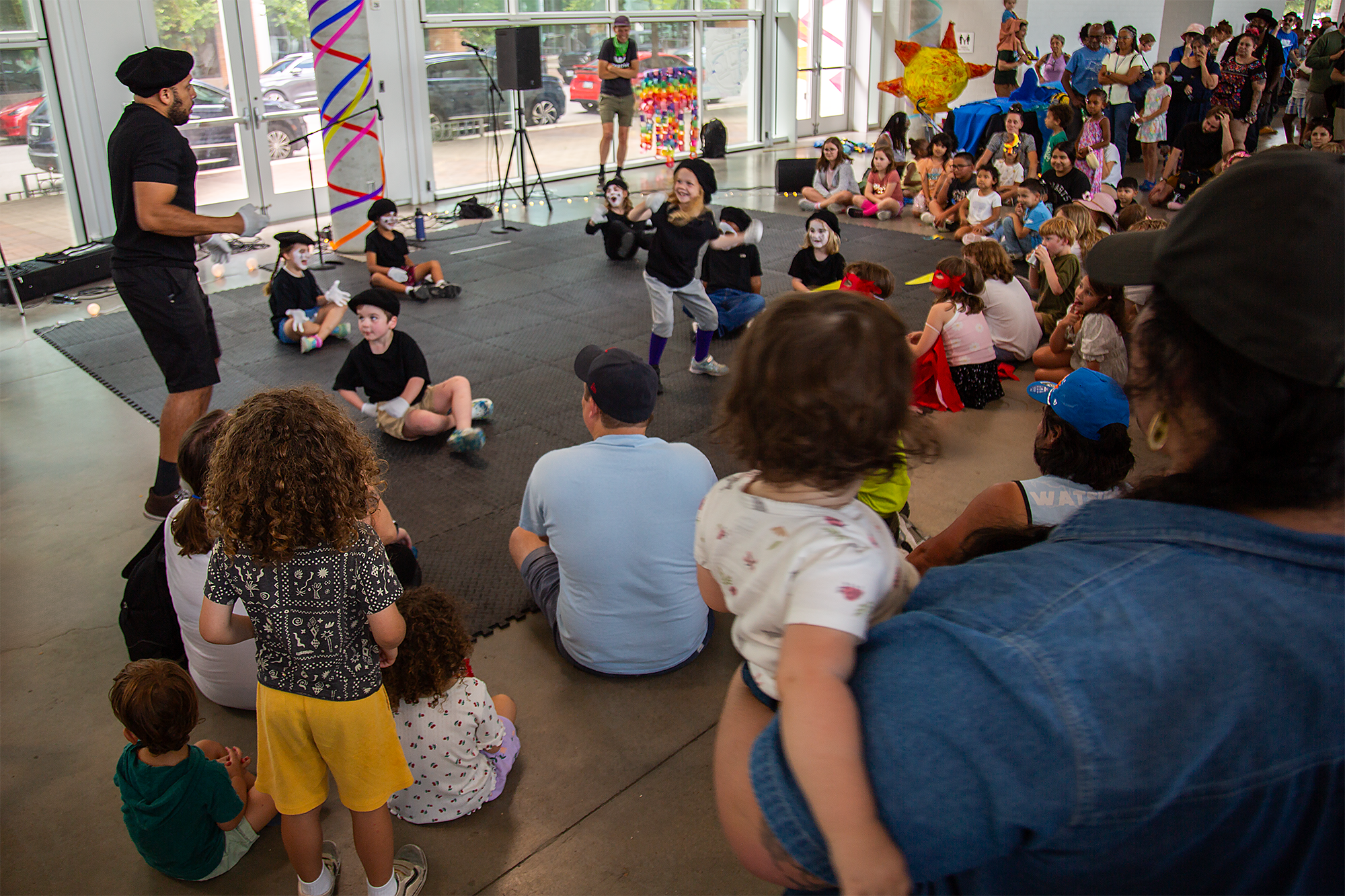 Children and adults gathered indoors at a children's event. Children are sitting on the floor, some wearing costumes or masks, with performers dressed as clowns or entertainers in front. The crowd watches the performance, which features colorful deco