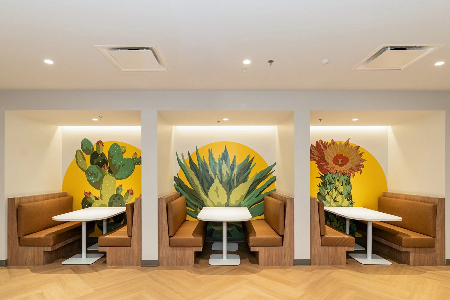 Three booth seating areas with brown cushions and white tables, each set against a wall with large colorful murals of cactus plants, including prickly pear, agave, and blooming cactus, on yellow background.