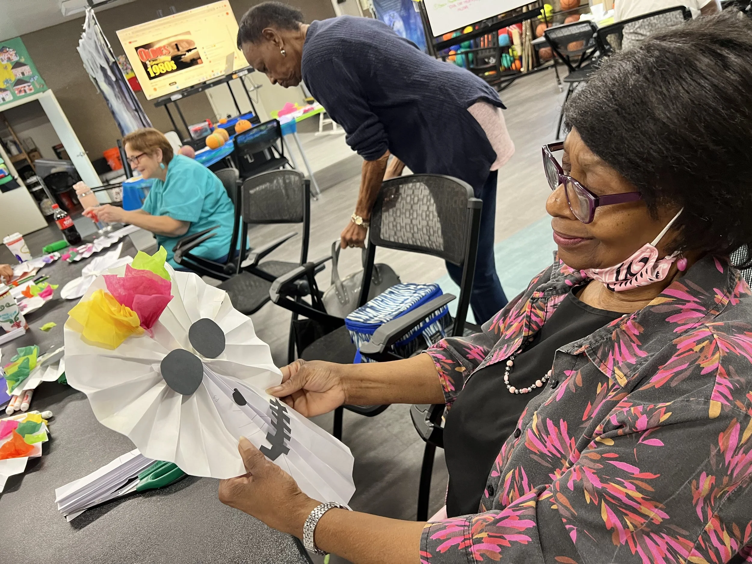A woman with glasses and a colorful floral blouse holds a large white poster or paper with black dots, sitting at a table with colorful paper decorations. Another woman in a turquoise shirt and a person in an orange shirt are seated nearby in a room decorated with colorful materials and a yellow sign in the background.