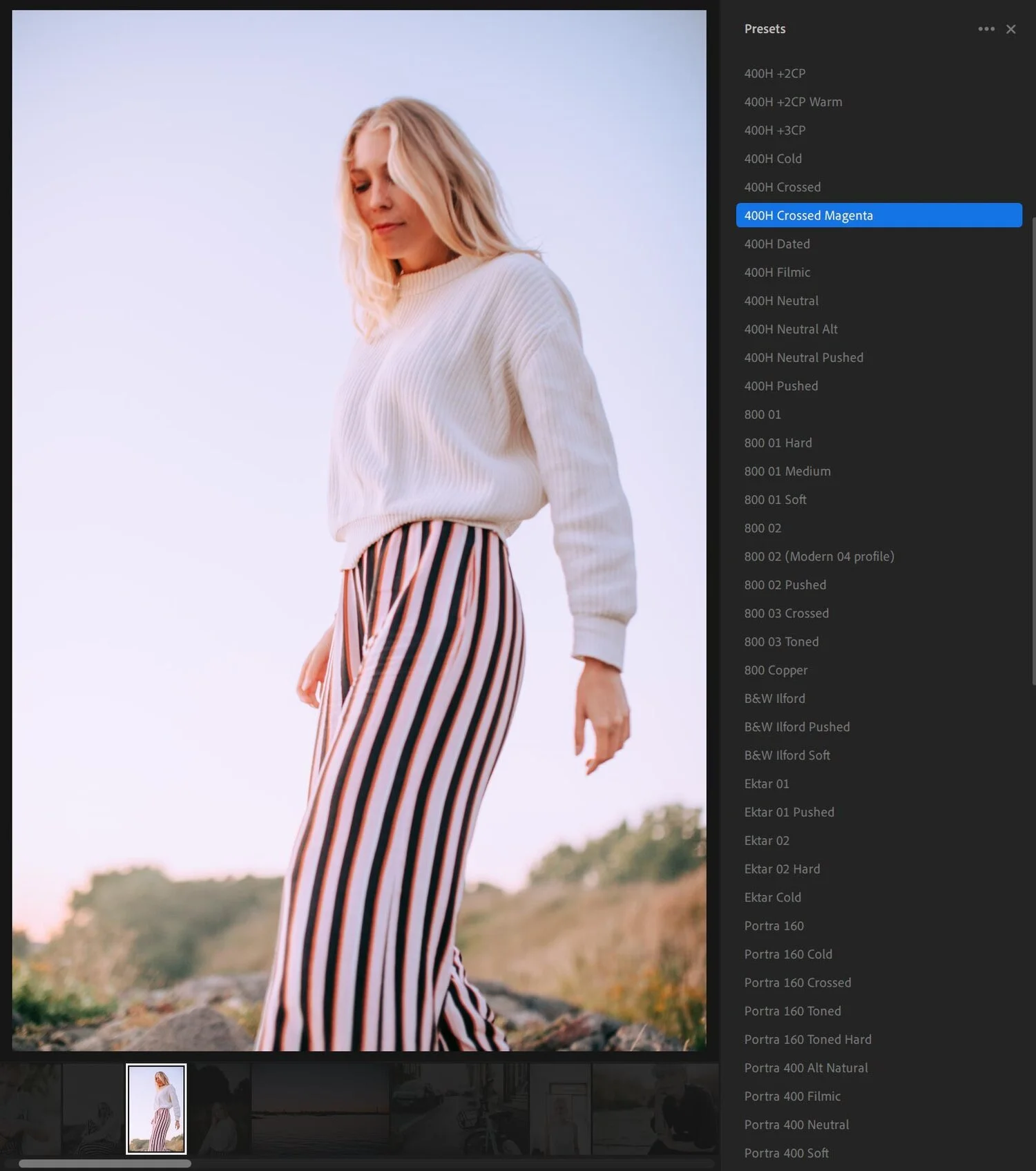 A woman with blonde hair wearing a white sweater and striped pants standing outdoors during sunset or sunrise.