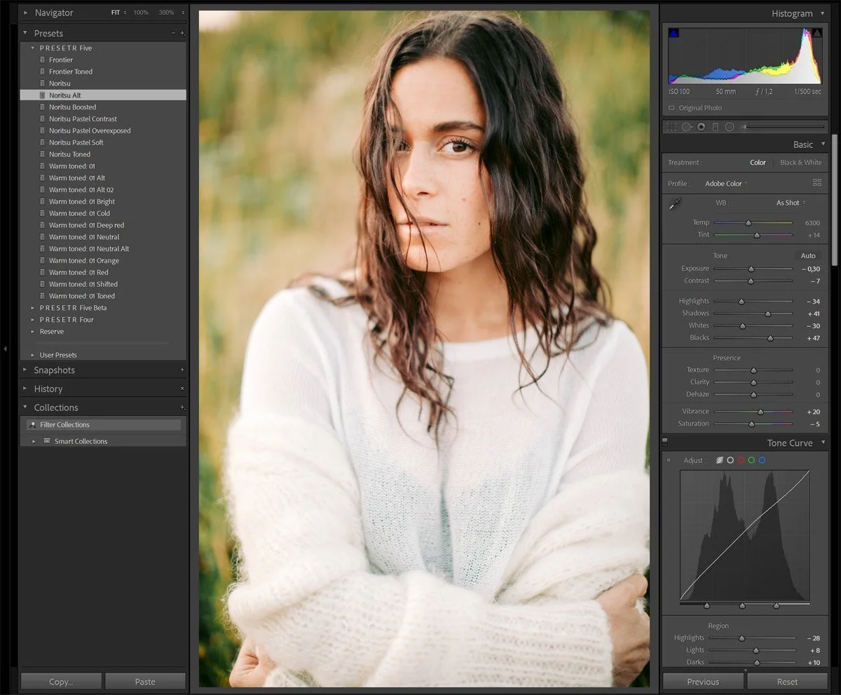 A young woman with wavy dark hair wearing a white sweater, standing outdoors with blurry greenery in the background.