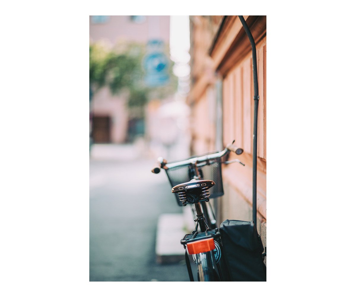 Close-up of an e-bike with a black basket, parked against an orange wooden wall on a city sidewalk, with blurred buildings and trees in the background.