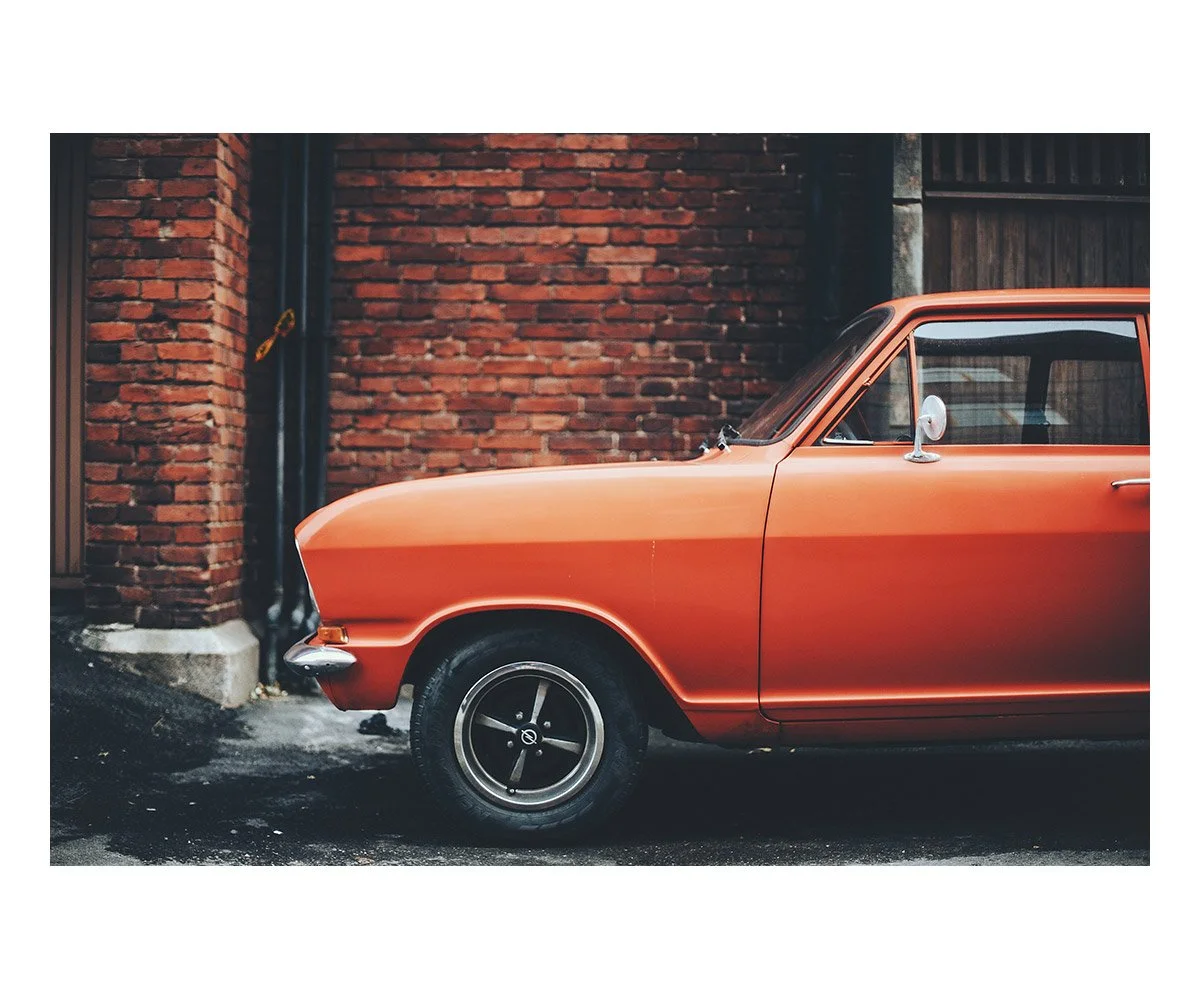 A vintage orange car parked on a street near a red brick wall.