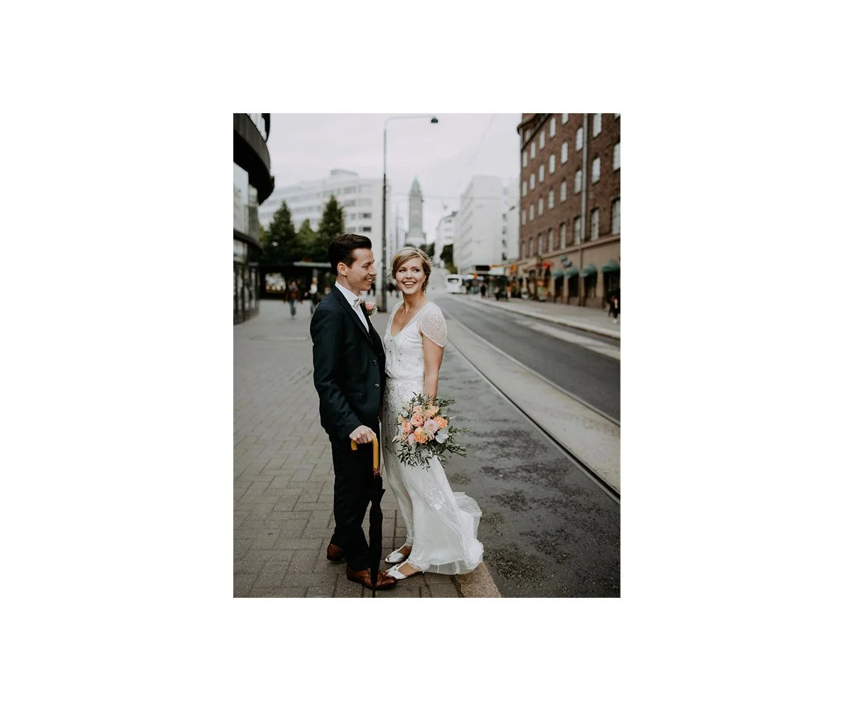 A newlywed couple standing on a city sidewalk, smiling at each other, with tram tracks and city buildings in the background. The bride is holding a bouquet of flowers.