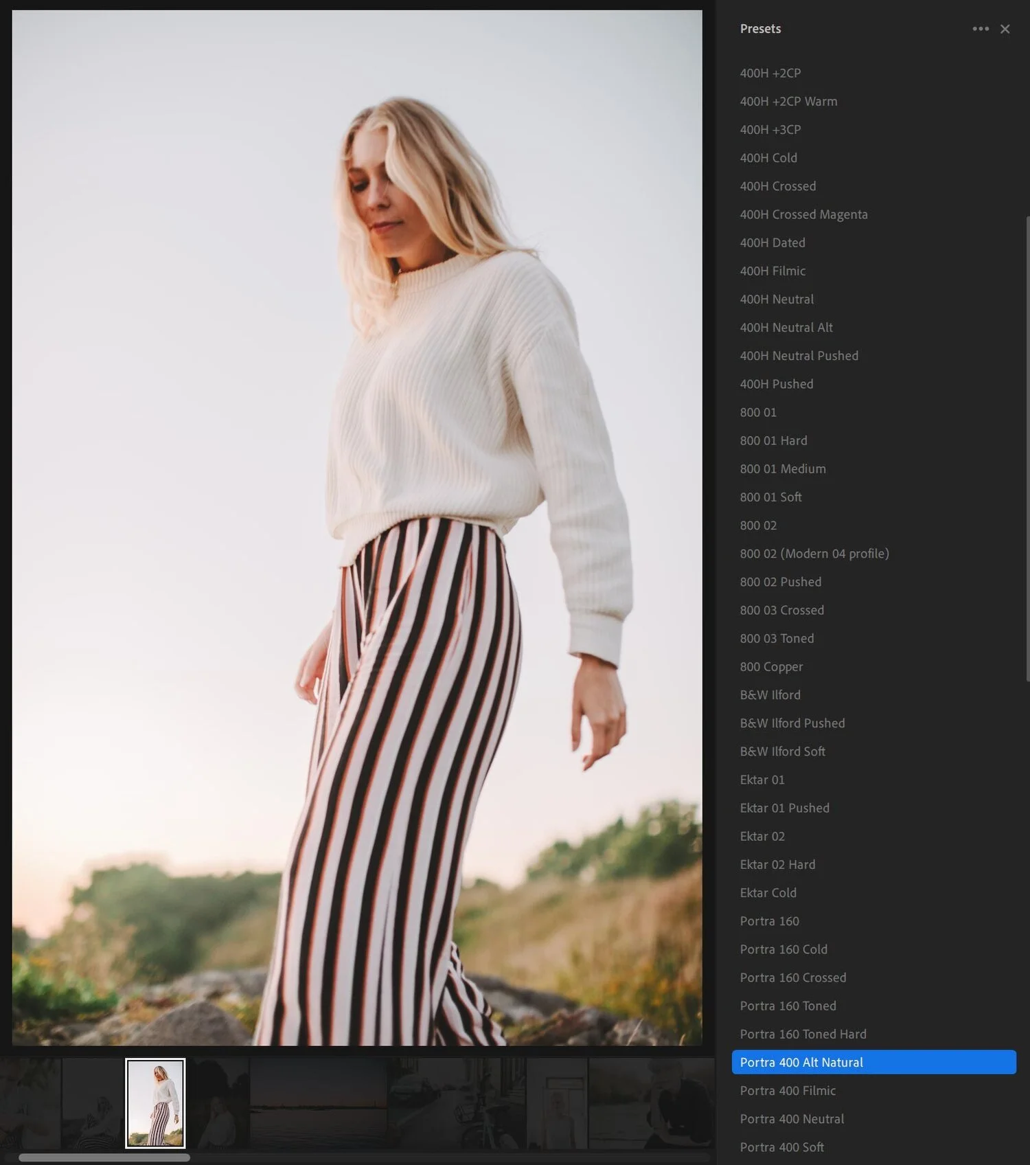 A woman with blonde hair wearing a white sweater and striped pants standing outdoors during sunset with a blurred natural background.