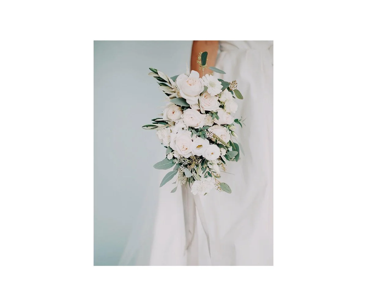 A person in a white dress holding a cascading bouquet of white roses, greenery, and other white flowers.
