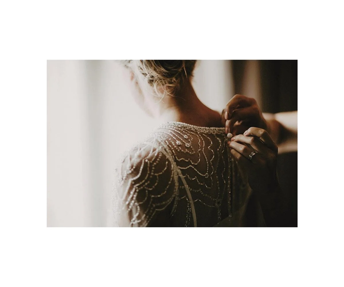 A woman in an embellished dress is adjusting a necklace or earring, with soft lighting creating a warm, intimate atmosphere.