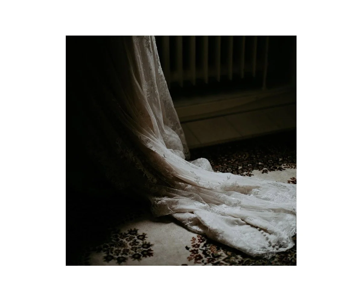A wedding dress on a rug, with a faint ribbed heater or radiator in the background, near a window with curtains.