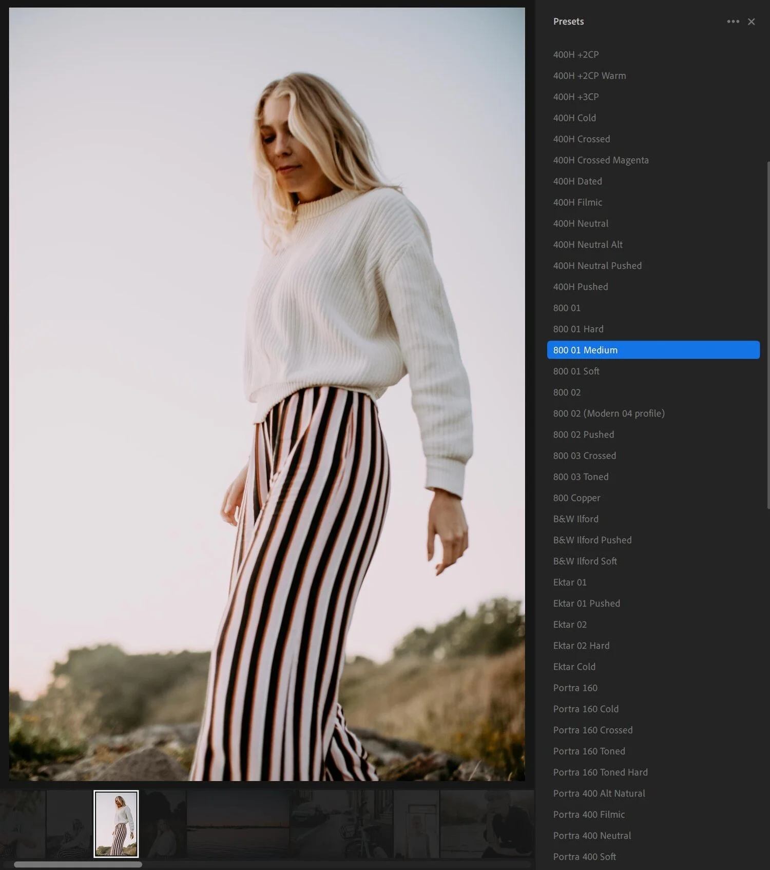 A woman in a white sweater and striped pants standing outdoors during sunset with a blurred natural landscape in the background.