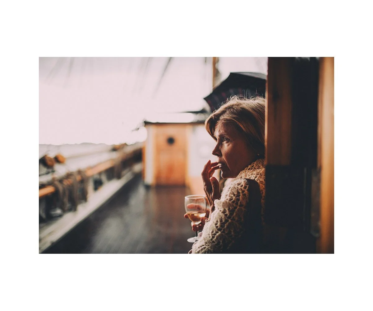 A woman sitting on a balcony or porch, holding a glass of wine, wearing a lace sweater, with a focused expression, overlooking an outdoor scene with some wooden structures and a blurred background.