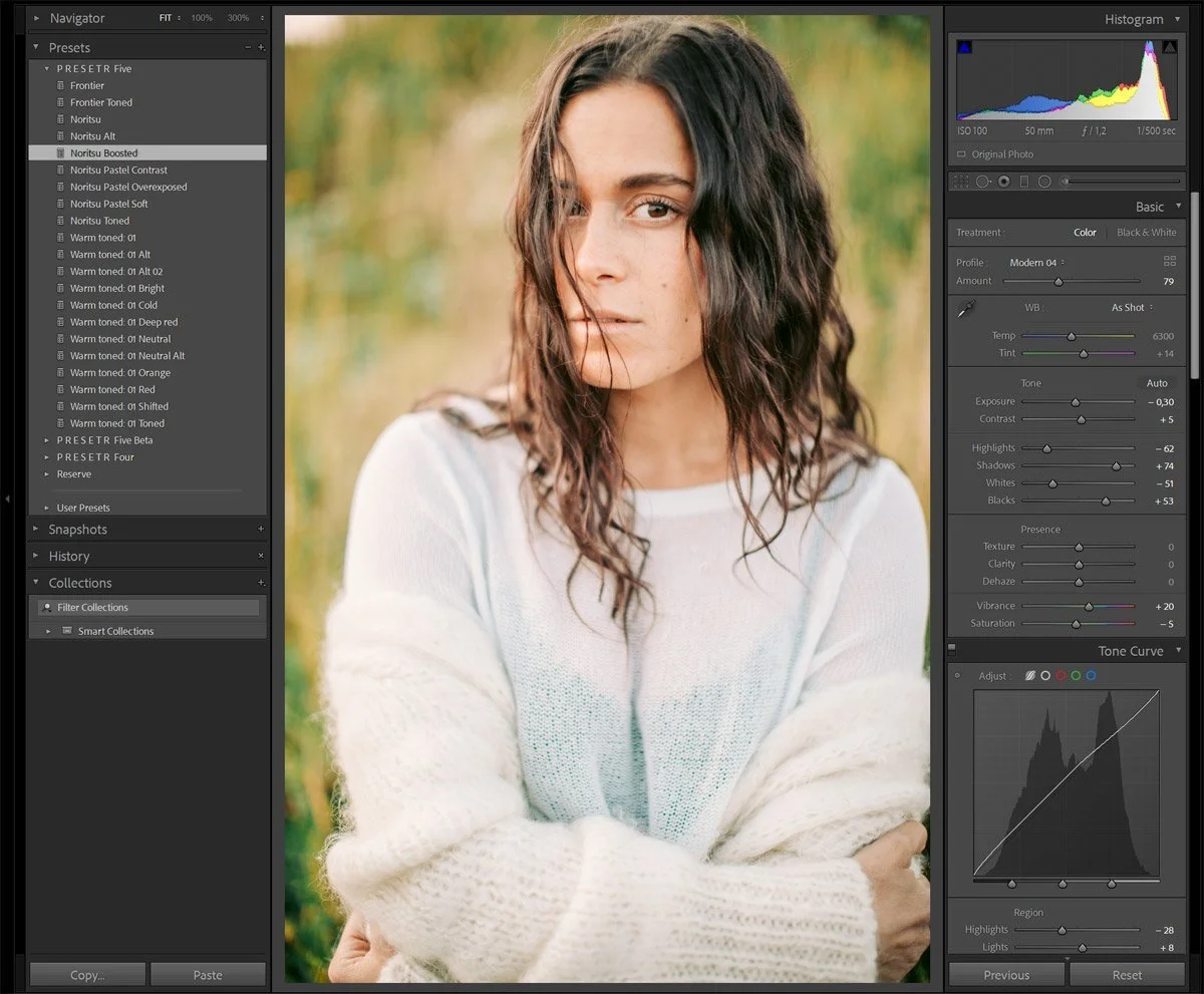 A young woman with wavy brown hair wearing a light-colored sweater, standing outdoors with blurred greenery in the background.
