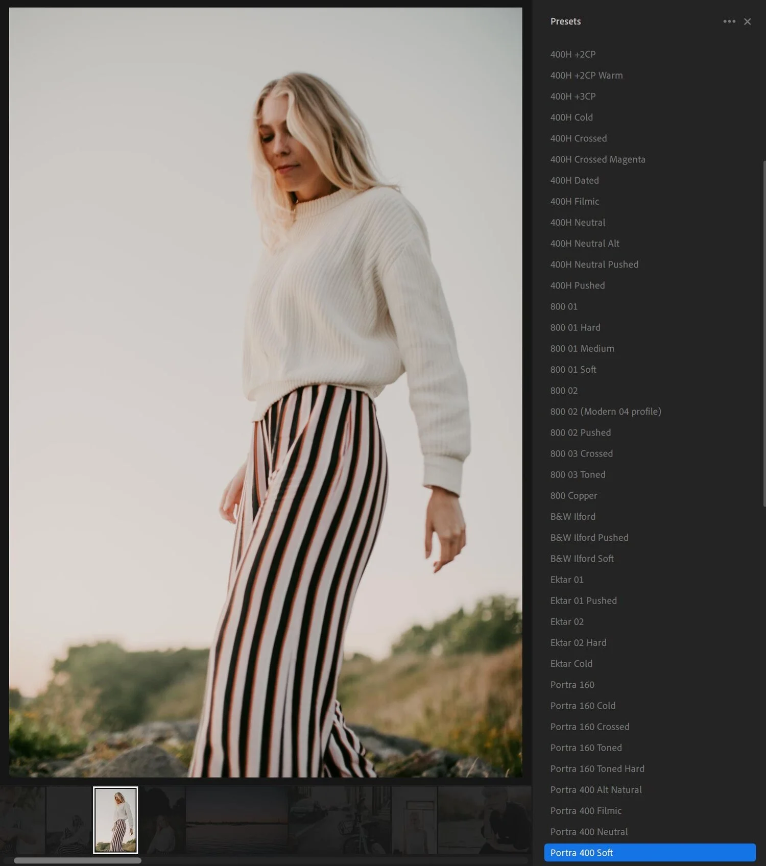 A young woman with blonde hair wearing a cream-colored sweater and striped pants standing outdoors.
