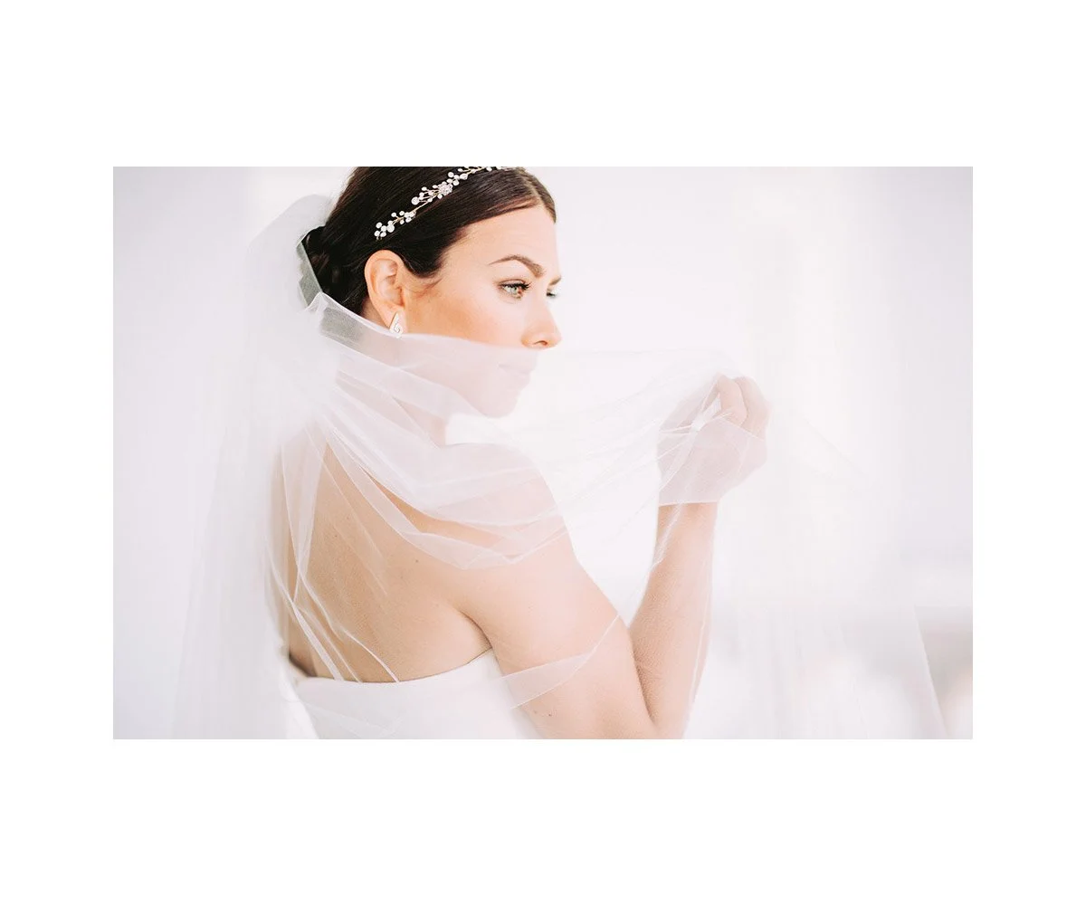 Bride in wedding dress with veil, looking sideways, holding her veil with one hand.
