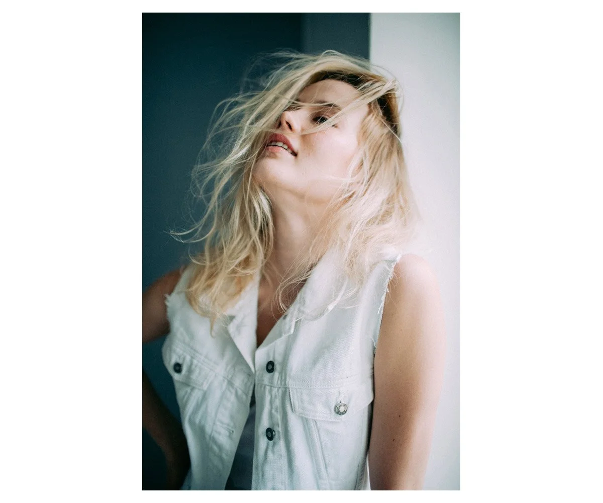 A young woman with blonde hair, wearing a sleeveless white denim vest, stands against a wall with her hair partially covering her face, looking slightly to the side with a relaxed and confident expression.
