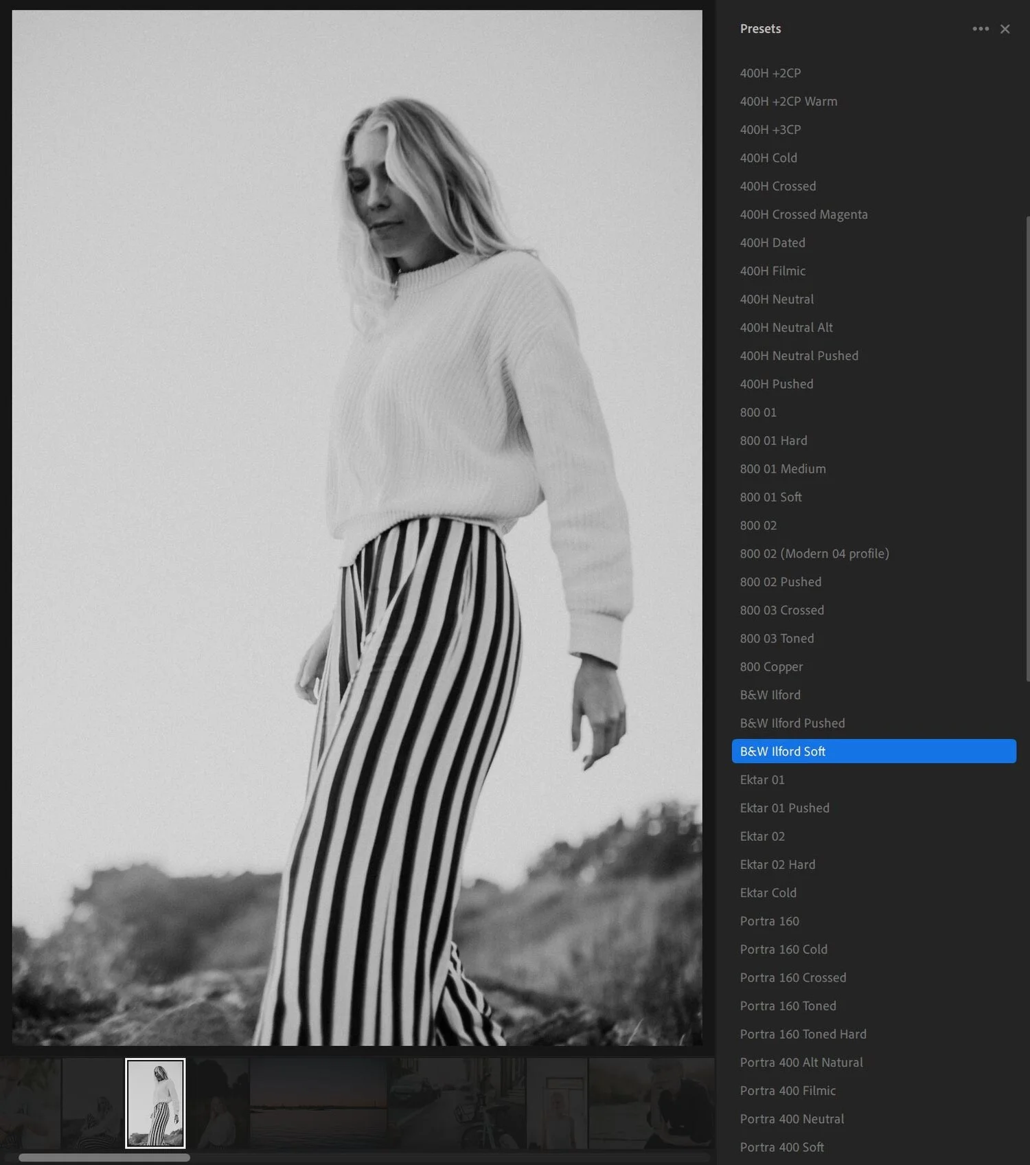 A woman with blonde hair wearing a light-colored sweater and high-waisted striped pants standing outdoors in a black-and-white photo.