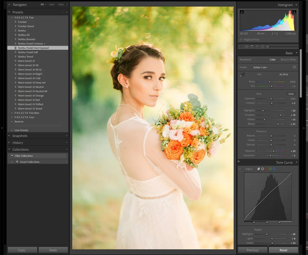 A woman in a wedding dress holding a colorful bouquet of flowers outdoors with sunlight and greenery in the background.