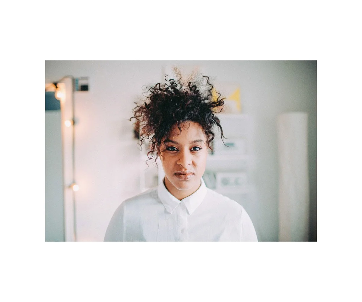 Portrait of a young woman with curly hair and a white shirt in a well-lit room.