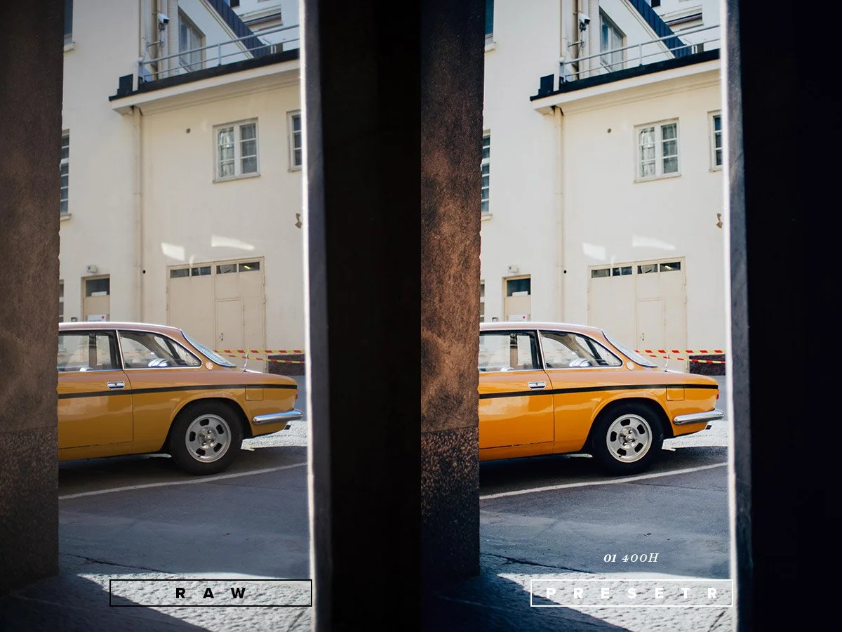 Photograph of a vintage yellow car parked on the street viewed through a narrow opening between two dark walls, with a beige building in the background.