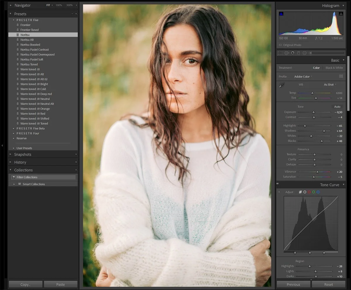 A woman with wavy dark hair and light skin wearing a white knit sweater, standing outdoors with a blurred natural background