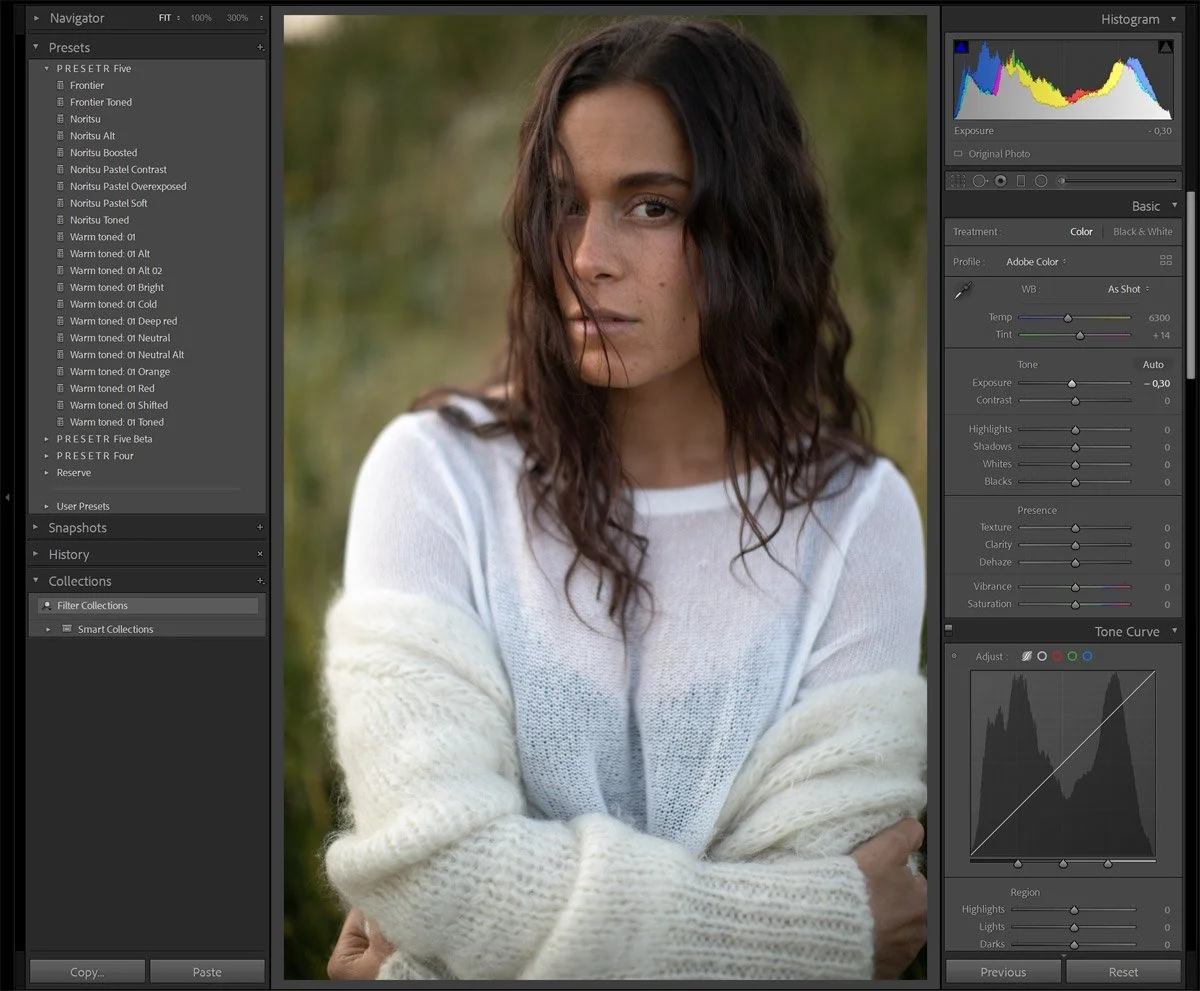 A young woman with wavy dark hair and light skin stands outdoors with blurred greenery in the background, wearing a white sweater and a partly sheer top.