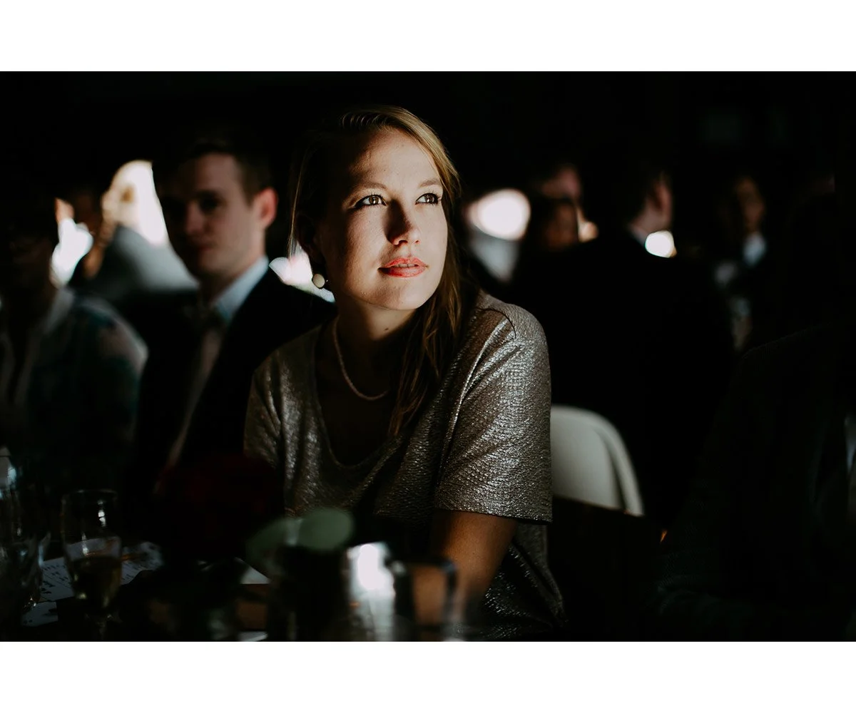 A woman with light skin and red hair attending a formal event, seated among other people in a dimly lit room. She is wearing a silver dress and pearl necklace, looking to her right with an attentive expression.