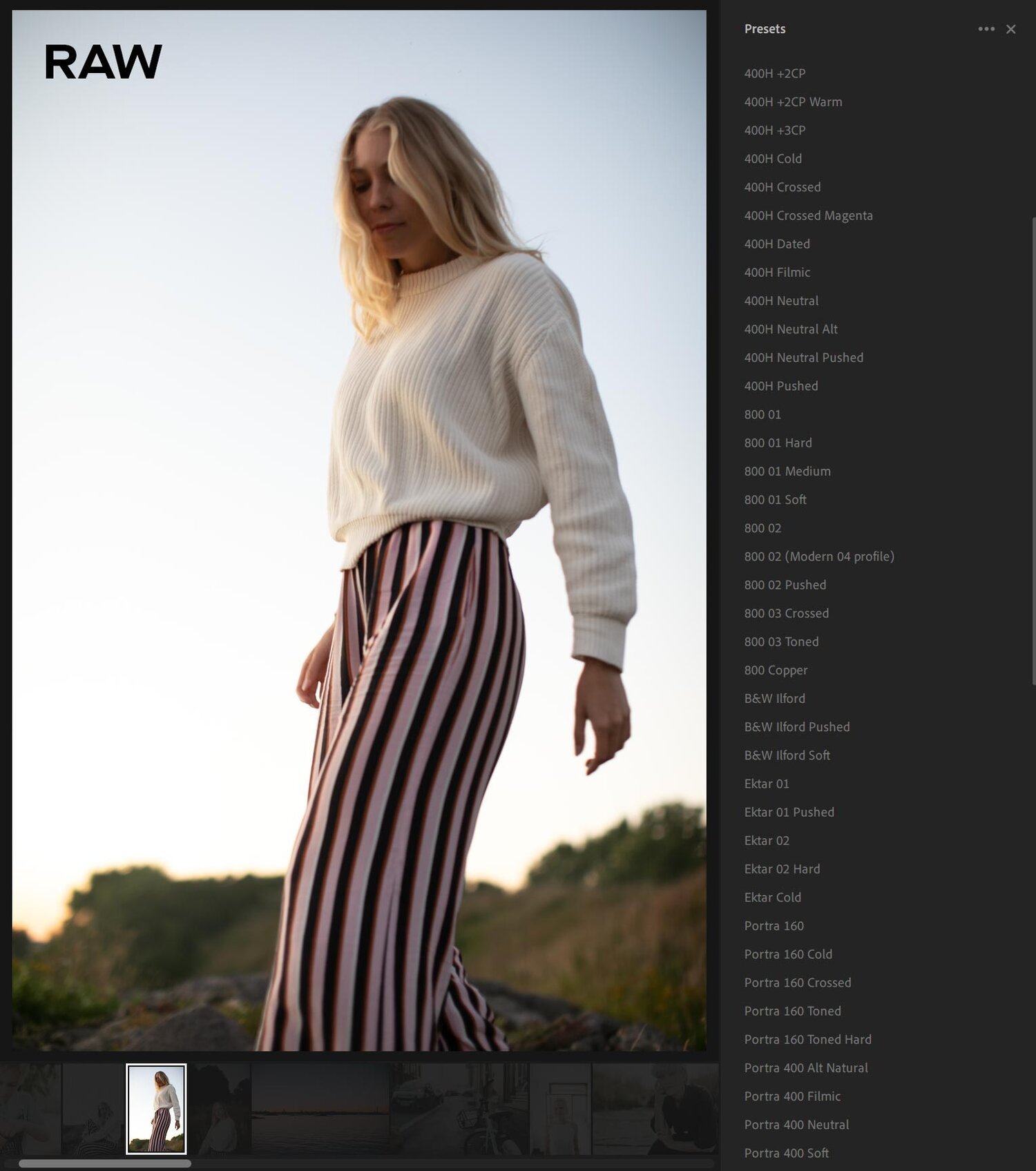A woman in a cream sweater and striped pants stands outdoors during sunset, with a clear sky and trees in the background.