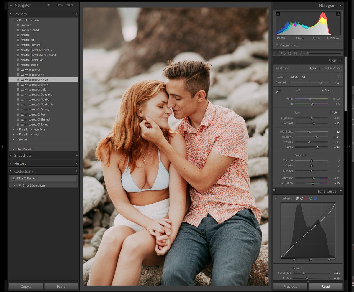A woman and man sit closely on rocks by the water, gazing at each other affectionately, with the woman resting her head gently on the man's forehead and their hands intertwined.