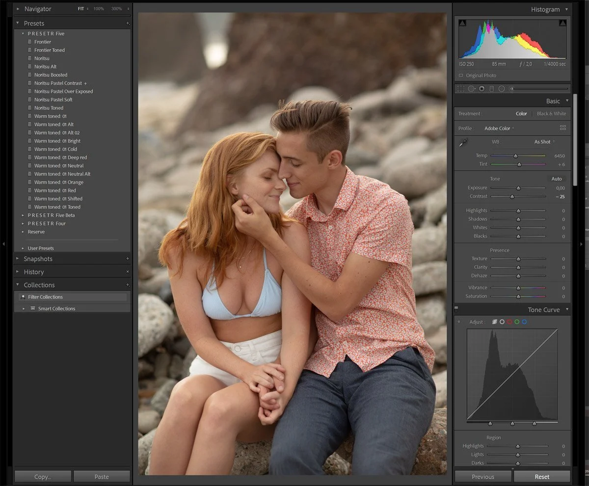 A young couple sitting together on rocks by a beach, with the woman wearing a light blue bikini top and white shorts, and the man in a short-sleeved floral shirt and dark pants, holding hands and leaning into each other with their foreheads touching.