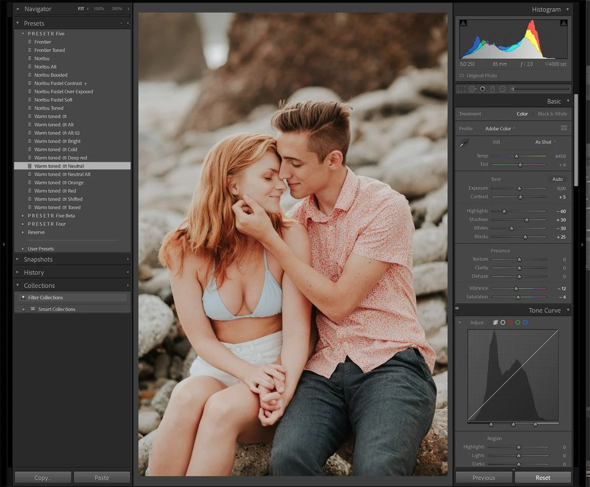 A young man and woman sitting close together on rocks near a body of water, sharing an intimate moment with their foreheads touching and eyes closed, surrounded by a rocky natural setting.