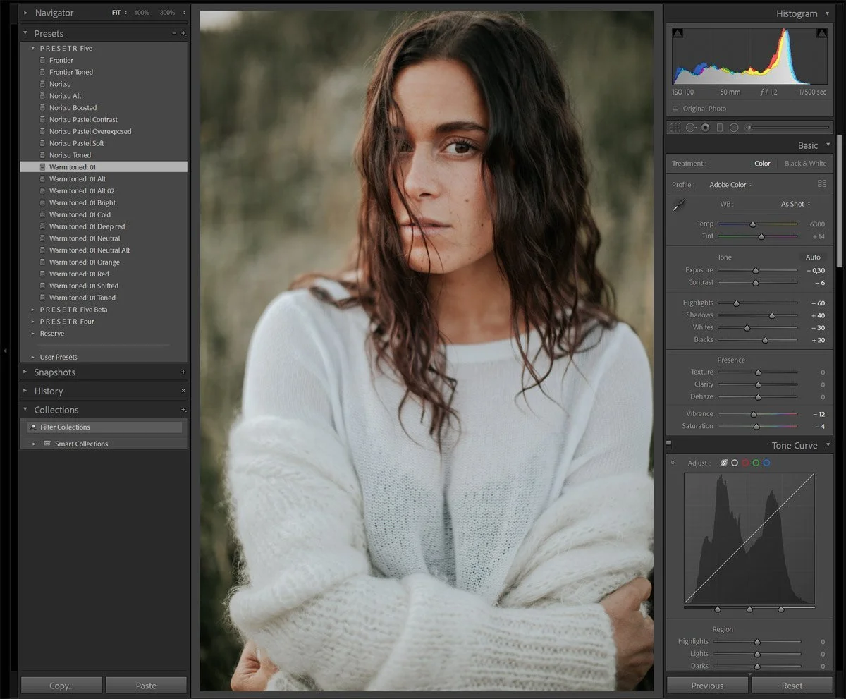 A woman with curly brown hair and freckles looks at the camera while wearing a white sweater in an outdoor setting with blurred greenery in the background.