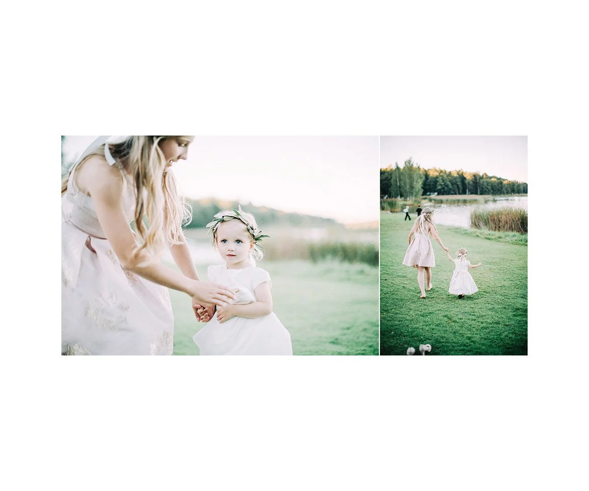 A woman and a young girl holding hands outdoors near a lake, with the woman leading and the girl following, both wearing light-colored dresses in a grassy area.