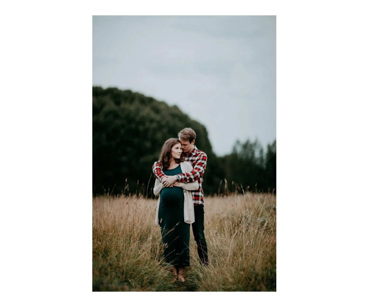 A couple standing in a grassy field, embracing each other, with a green hill and trees in the background.