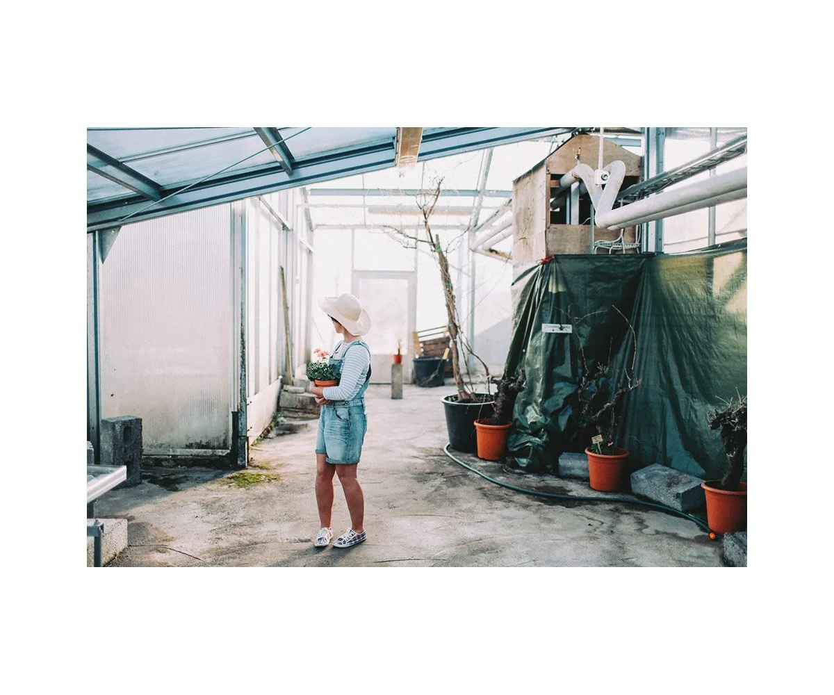 A girl wearing a sunhat, striped shirt, and denim overalls in a greenhouse, holding a pot of flowers.