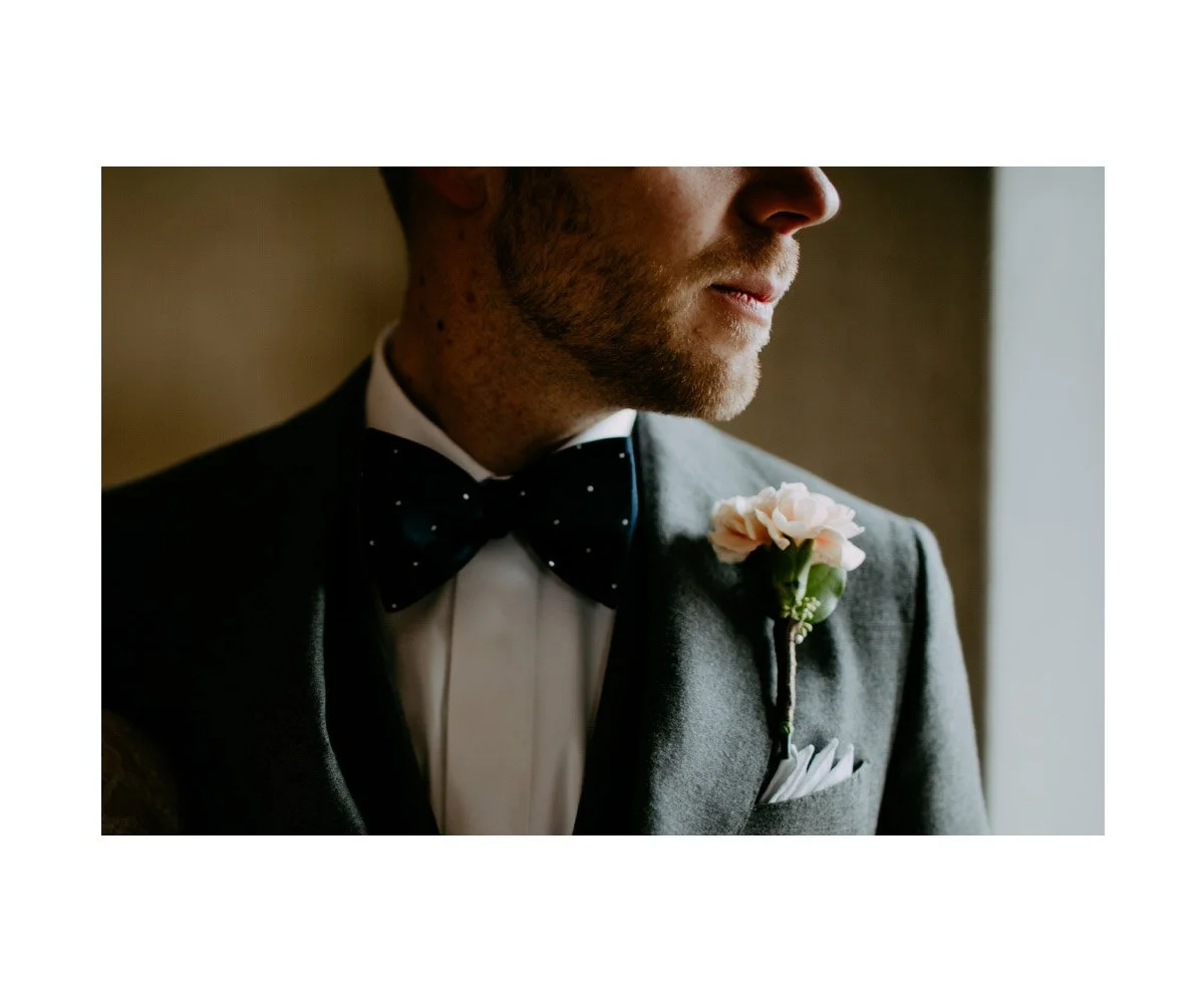 Close-up of a groom in a tuxedo with a black bow tie and a pink boutonniere, looking to the side by a window.