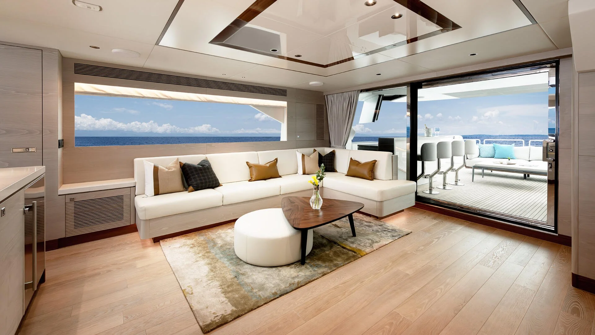 Luxury yacht interior with a white sectional sofa, a wooden coffee table with a vase of flowers, and a sliding glass door leading to a deck with ocean views.