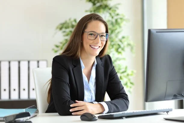A woman with glasses sitting at a desk in an office, smiling at the camera. She is wearing a black blazer and white shirt, with a computer monitor in front of her and a green plant in the background.