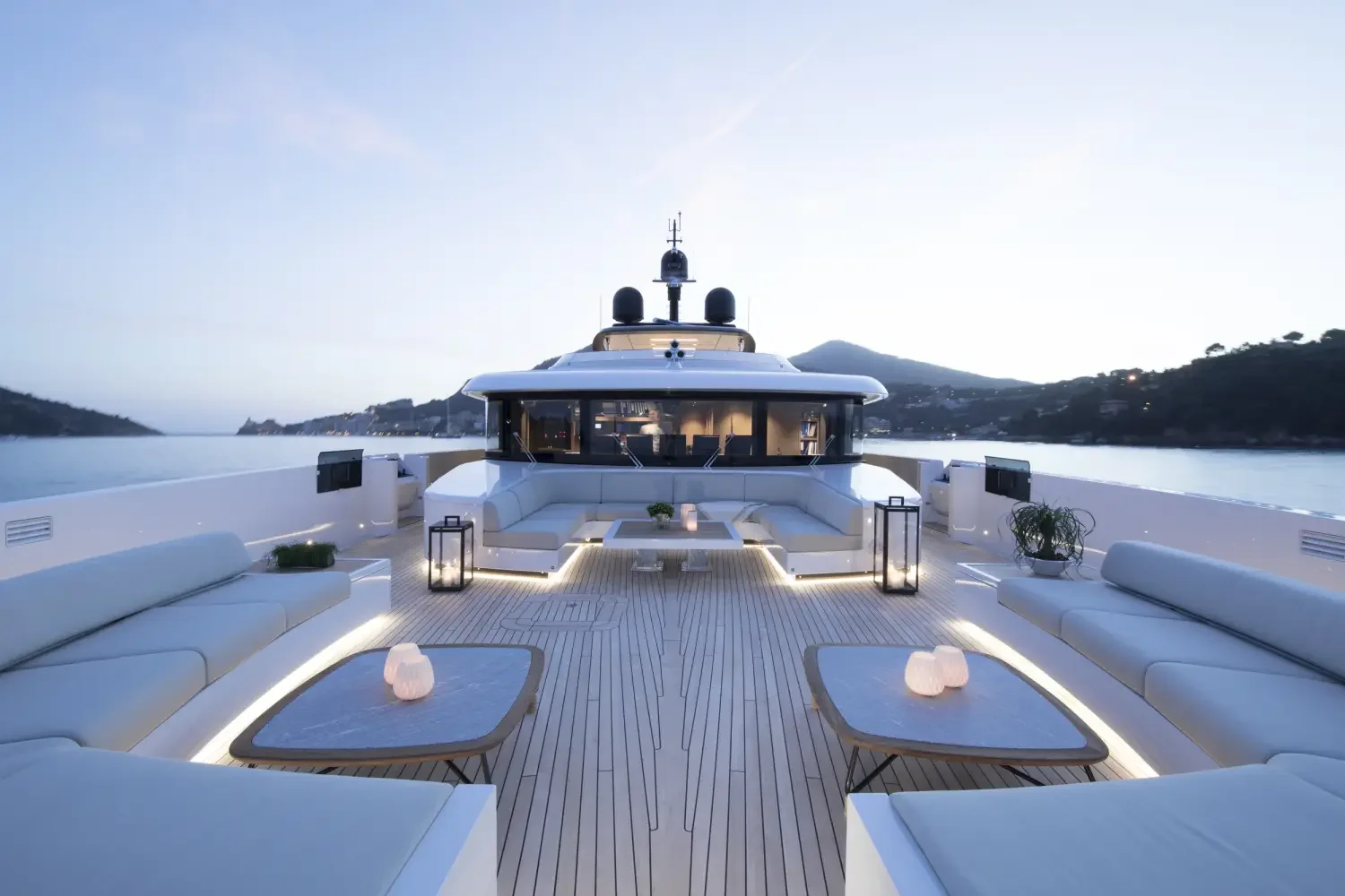Luxury yacht deck with white seating, tables, and lanterns, overlooking calm water with mountainous shoreline in the background during dusk.