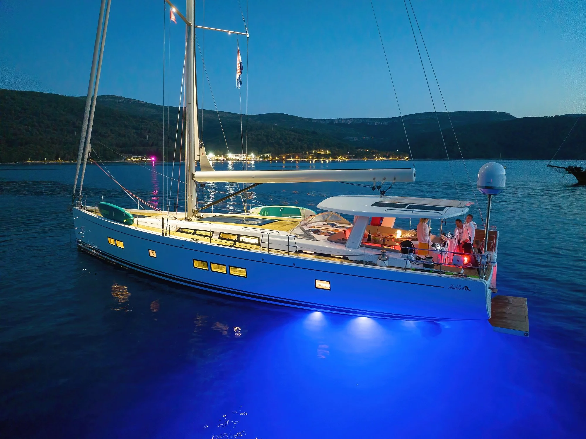 A luxury yacht sailing on calm waters at night with a group of people gathering on the deck, illuminated by colorful lights, with a mountain and city lights visible in the background.