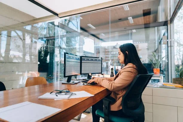 A woman working at a desk with two computer monitors in a modern office with glass walls.