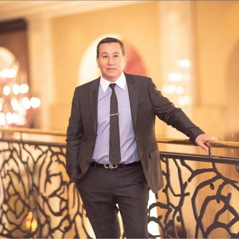 A man in a black suit and tie standing on a balcony with a decorative black railing, in a warmly lit, elegant interior setting.