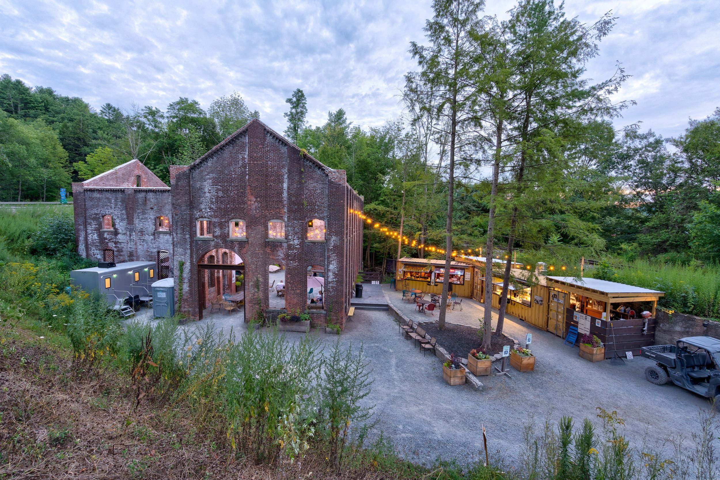 Outdoor dining area with a rustic brick building and food stalls surrounded by trees, with string lights illuminating the setting.